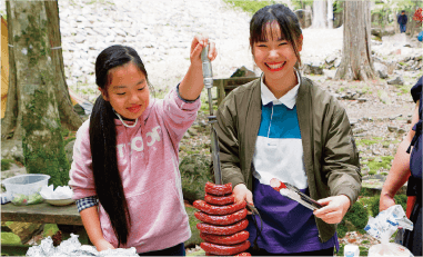 鉄串に刺したシュラスコスタイルで焼き上げた鹿肉フランクをカットする薬師神杏美(やくしじんあみ)さん(左)と田中さん