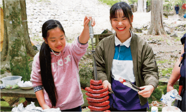 鉄串に刺したシュラスコスタイルで焼き上げた鹿肉フランクをカットする薬師神杏美（やくしじんあみ）さん（左）と田中さん